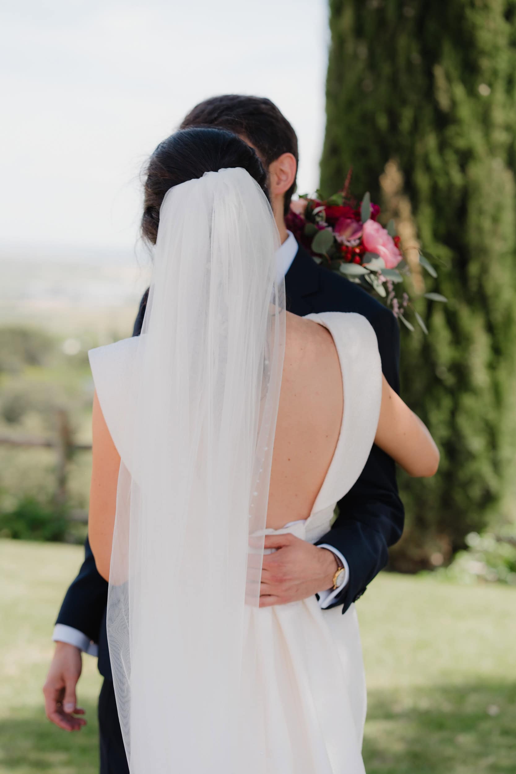 Bride and groom embracing with flowing veil and green bouquet by Estudio Zoe